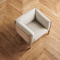 Beige upholstered chair with light wood frame on a herringbone-patterned wood floor. Modern, minimalist design.