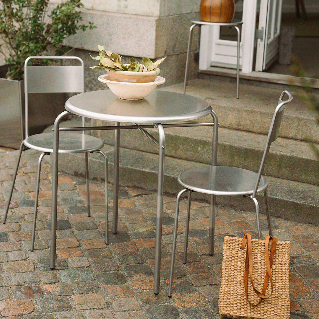 Silver metal table and chairs with a minimalistic design, set on a cobblestone patio with greenery backdrop.