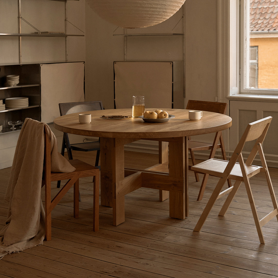 "Natural wood round dining table with light beige chairs and soft beige blanket in a warm-toned interior setting."