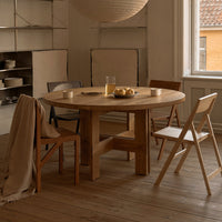 "Natural wood round dining table with light beige chairs and soft beige blanket in a warm-toned interior setting."