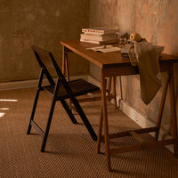 Wooden desk in warm tones with a black folding chair, neutral walls, and textured beige rug. Stylish and cozy workspace.
