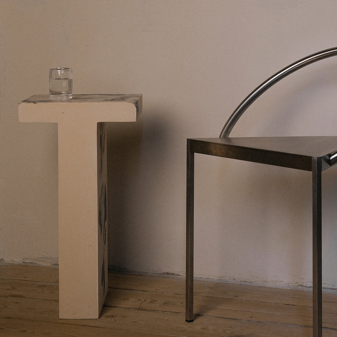 Modern beige table with cut-out patterns next to a sleek metallic chair, set against a neutral wall and wooden floor.