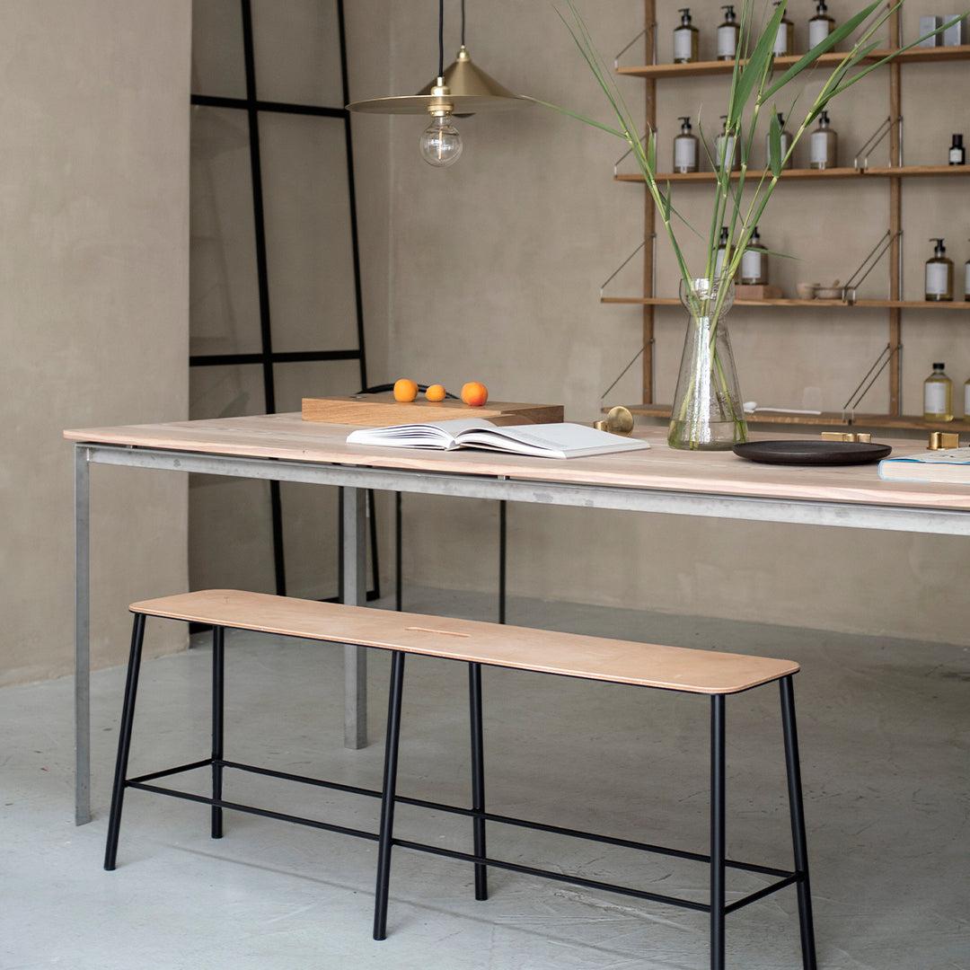 Light wood dining table with a smooth finish and black metal legs, complemented by a sleek black bench, set in a neutral-toned room.