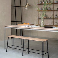 Light wood dining table with a smooth finish and black metal legs, complemented by a sleek black bench, set in a neutral-toned room.