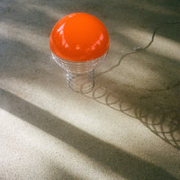 Glossy orange round lamp with a spiral metallic base casting soft shadows on a textured floor.