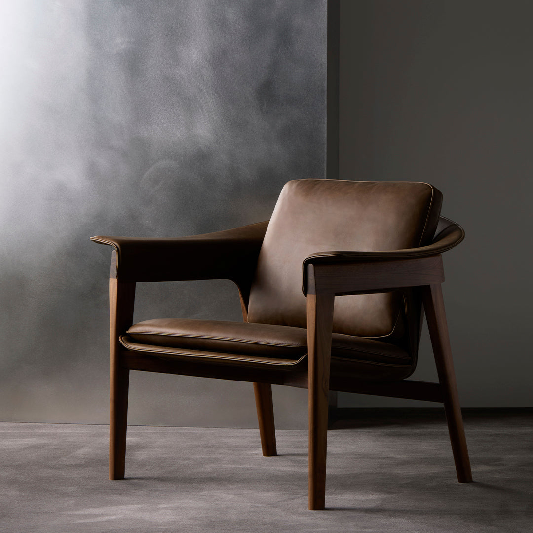 Brown leather armchair with a sleek wooden frame, featuring a minimalist design against a textured gray backdrop.