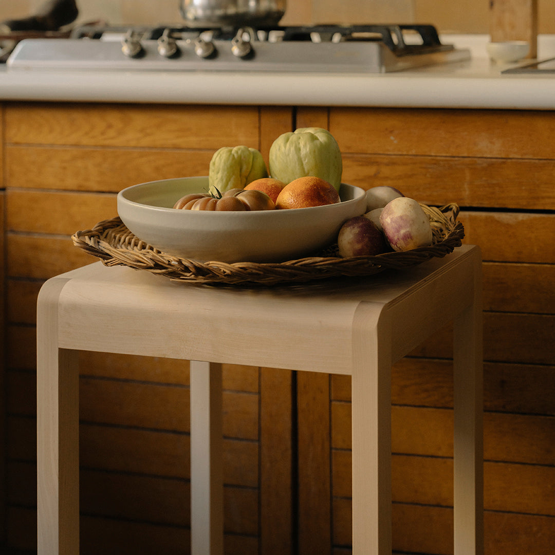 Light beige table with woven basket of colorful fruits against a warm wood kitchen backdrop. Natural tones, earthy aesthetic.