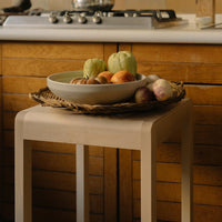 Light beige table with woven basket of colorful fruits against a warm wood kitchen backdrop. Natural tones, earthy aesthetic.