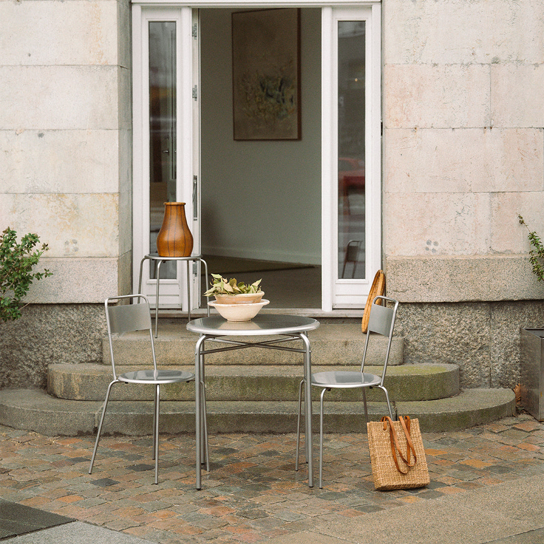 Outdoor table set featuring silver metal chairs, a round glass table, and a beige woven bag against textured stone steps.