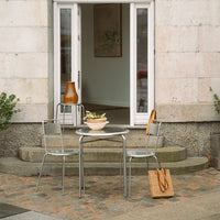 Outdoor table set featuring silver metal chairs, a round glass table, and a beige woven bag against textured stone steps.