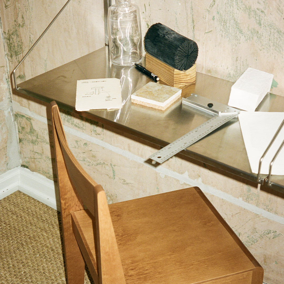 Wooden chair with a natural finish, metallic desk surface, glass and paper accessories in a textured beige and green background.