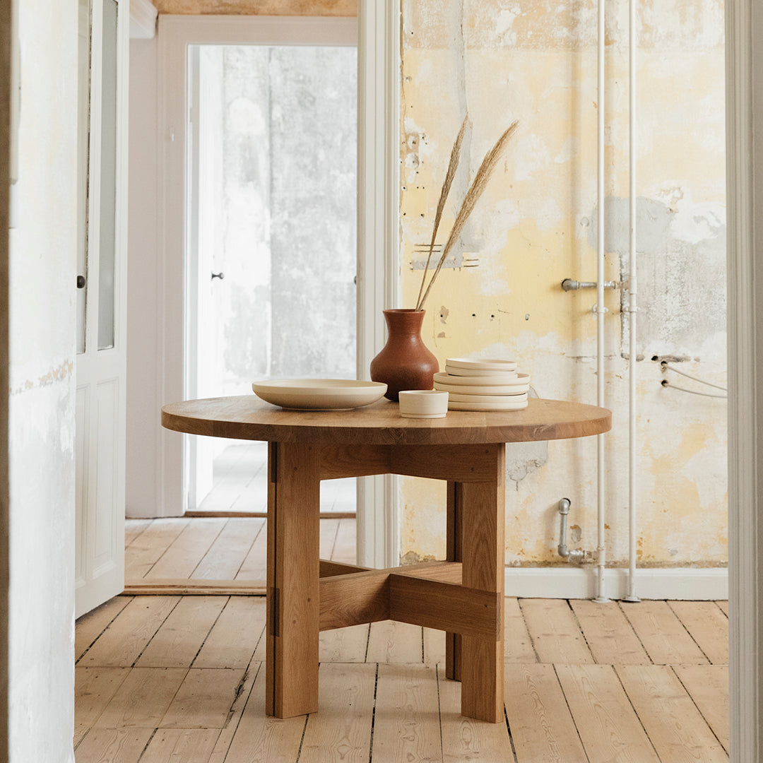 Round wooden table with a natural finish, complemented by a terracotta vase and neutral plate set.