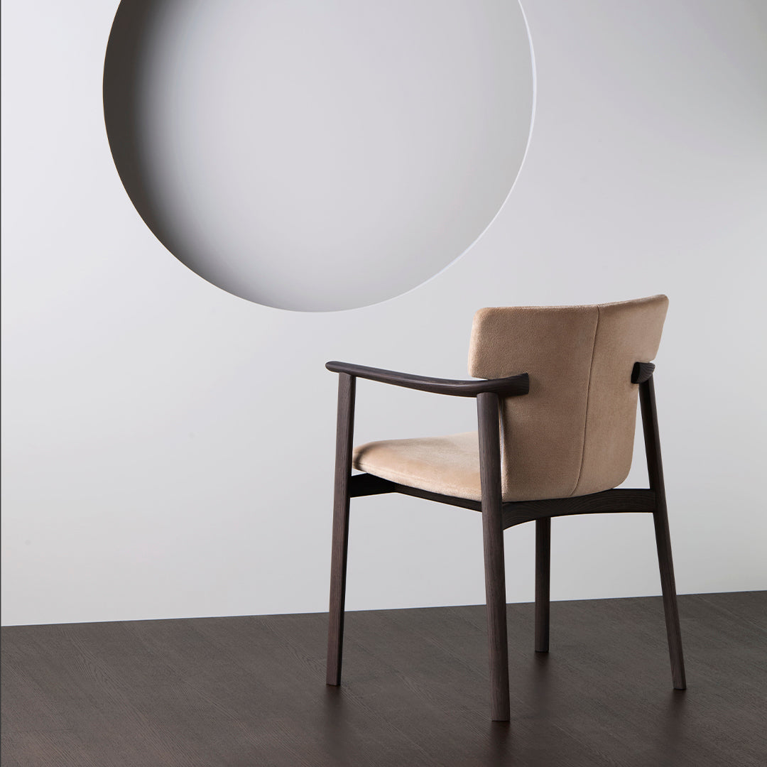 Beige upholstered chair with dark wooden frame, set against a minimalist wall with a circular cutout.