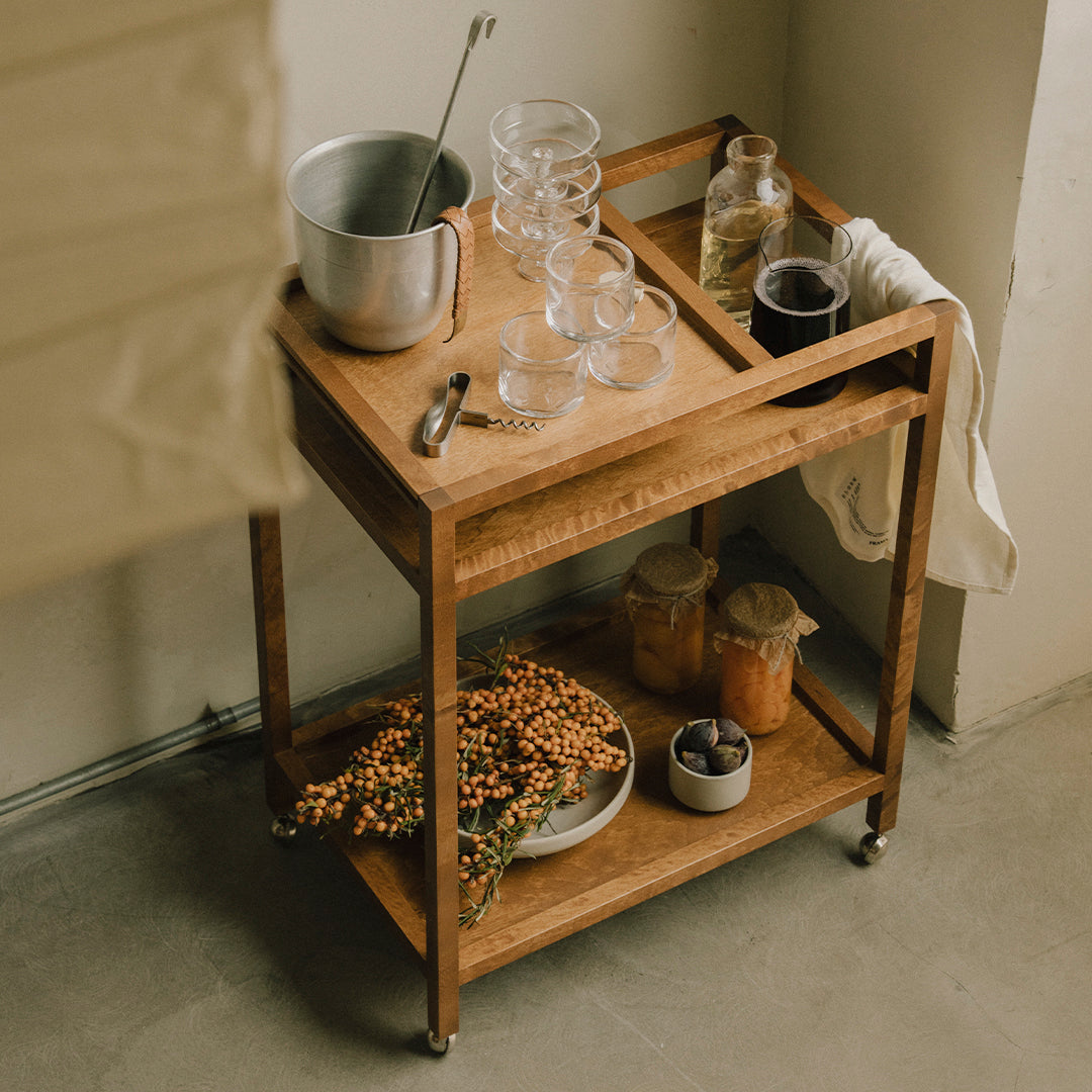 Wooden serving cart featuring natural tones and rustic decor, with clear glasses, metal accents, and earthy plant arrangement.
