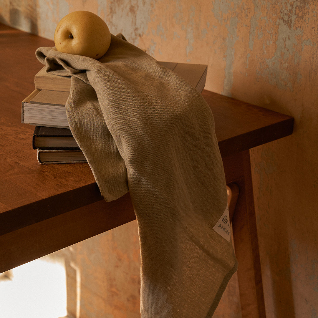 Beige linen towel draped over a wooden table, complemented by a yellow apple and stacked books.