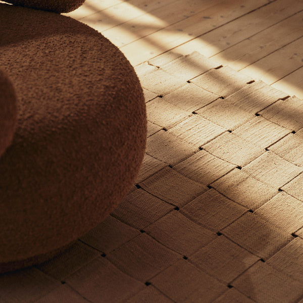 Brown textured pouf on a woven beige floor, accented by soft natural shadows and geometric patterns.