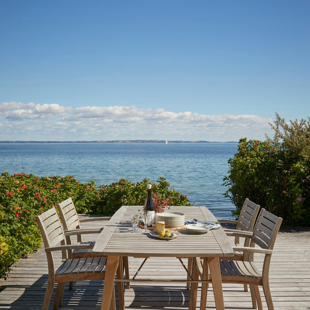 "Light wood dining set on a deck, featuring natural tones with a serene blue ocean backdrop and vibrant green foliage."