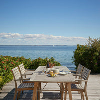 "Light wood dining set on a deck, featuring natural tones with a serene blue ocean backdrop and vibrant green foliage."