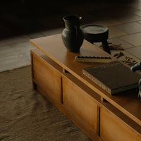 Wooden coffee table with a smooth, warm brown finish; styled with a black vase and stacked books. Textured beige rug beneath.