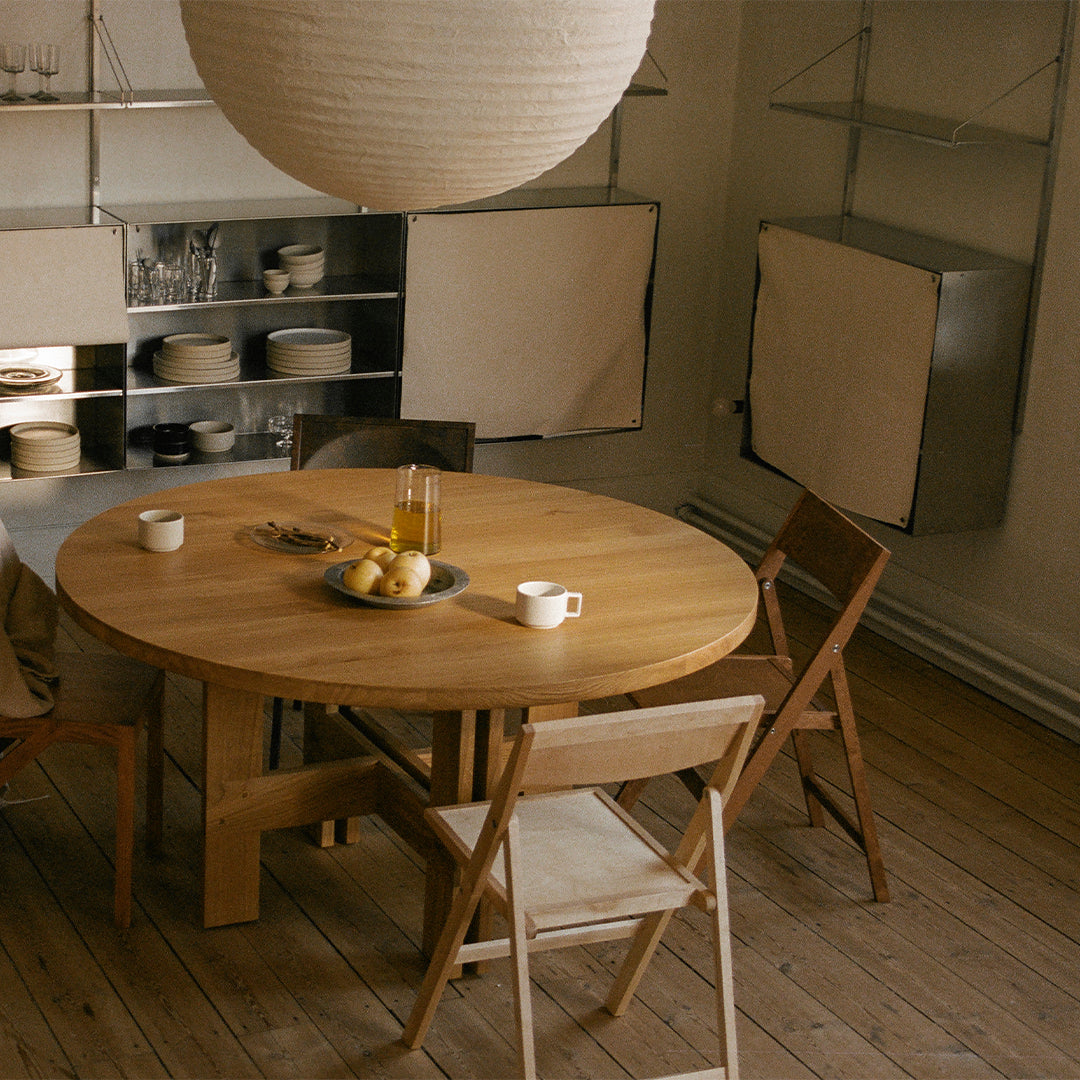 Natural wood round dining table with light beige folding chairs, soft white pendant lamp, and minimalist decor.