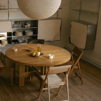 Natural wood round dining table with light beige folding chairs, soft white pendant lamp, and minimalist decor.