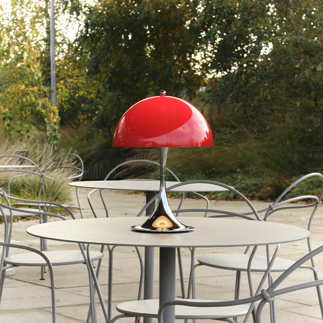 Red dome lamp with a sleek chrome base, placed on a modern gray tabletop surrounded by minimalist chairs.