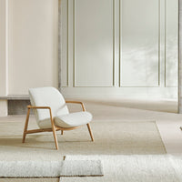 Light gray upholstered chair with a natural wood frame, set against a soft textured rug and neutral wall tones.