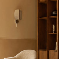 Sleek silver wall sconce against warm beige wall, complemented by natural wood shelving and textured cream chair.