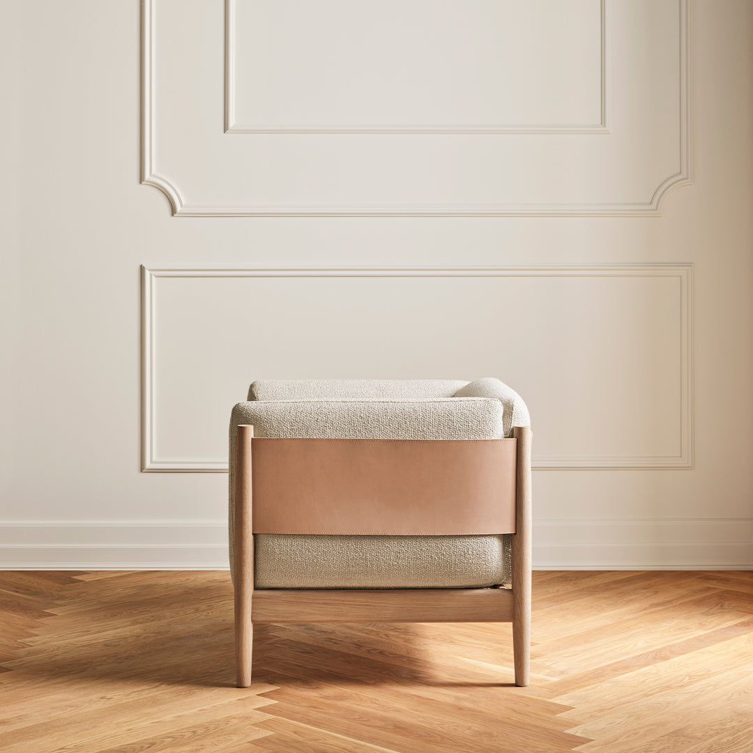 Beige upholstered chair with light wood frame and a smooth brown leather accent against a white wall.