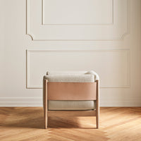 Beige upholstered chair with light wood frame and a smooth brown leather accent against a white wall.