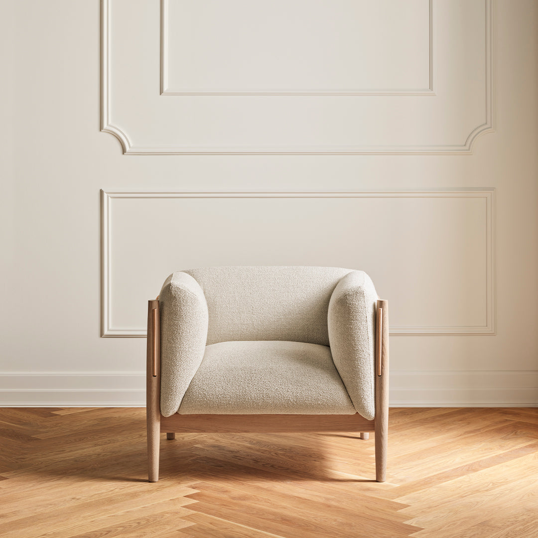 Cream-colored upholstered chair with rounded arms and light wood frame, set against a neutral wall and herringbone floor.