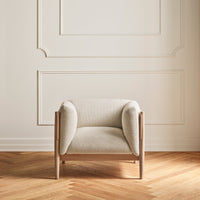 Cream-colored upholstered chair with rounded arms and light wood frame, set against a neutral wall and herringbone floor.