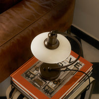 Bronze table lamp with a white shade on orange stacked books, set against a brown leather sofa and black-and-white patterned floor.