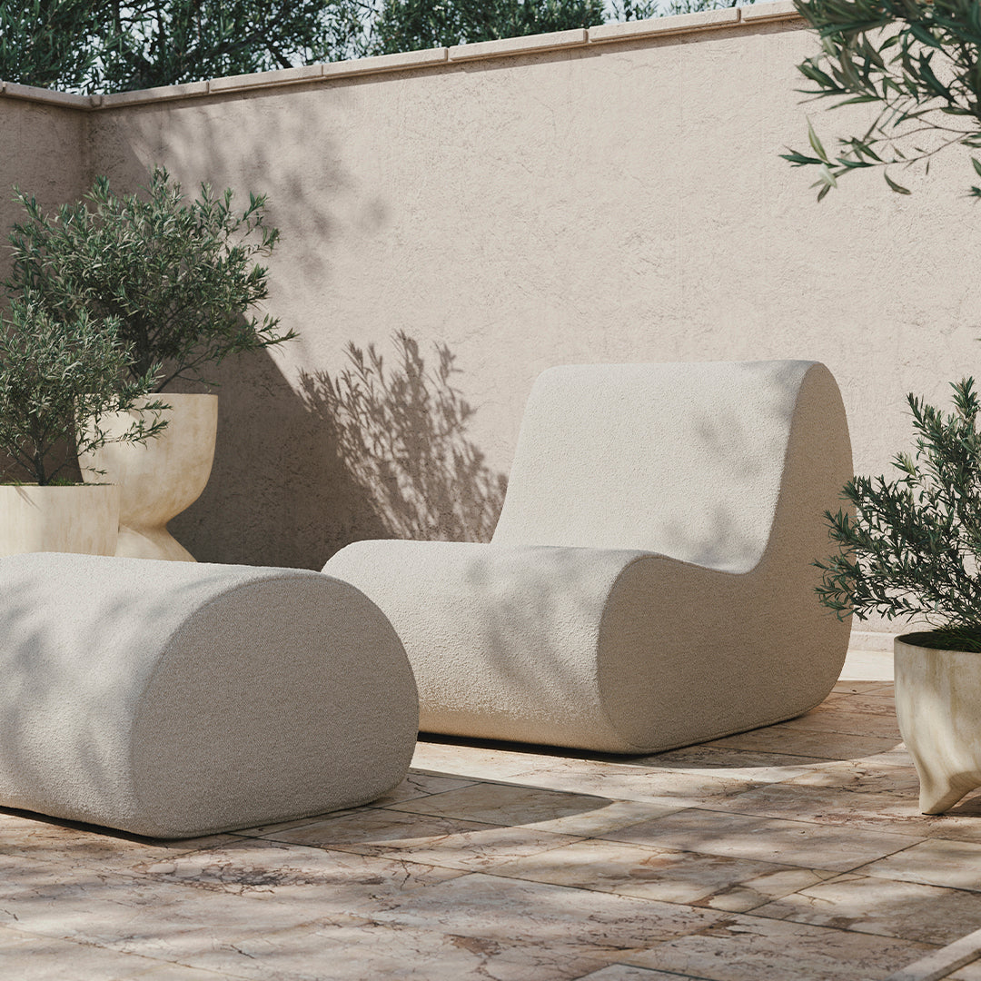 Beige sculptural lounge chair and ottoman with smooth curves, set against a textured light stone patio with green plants.