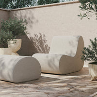 Beige sculptural lounge chair and ottoman with smooth curves, set against a textured light stone patio with green plants.