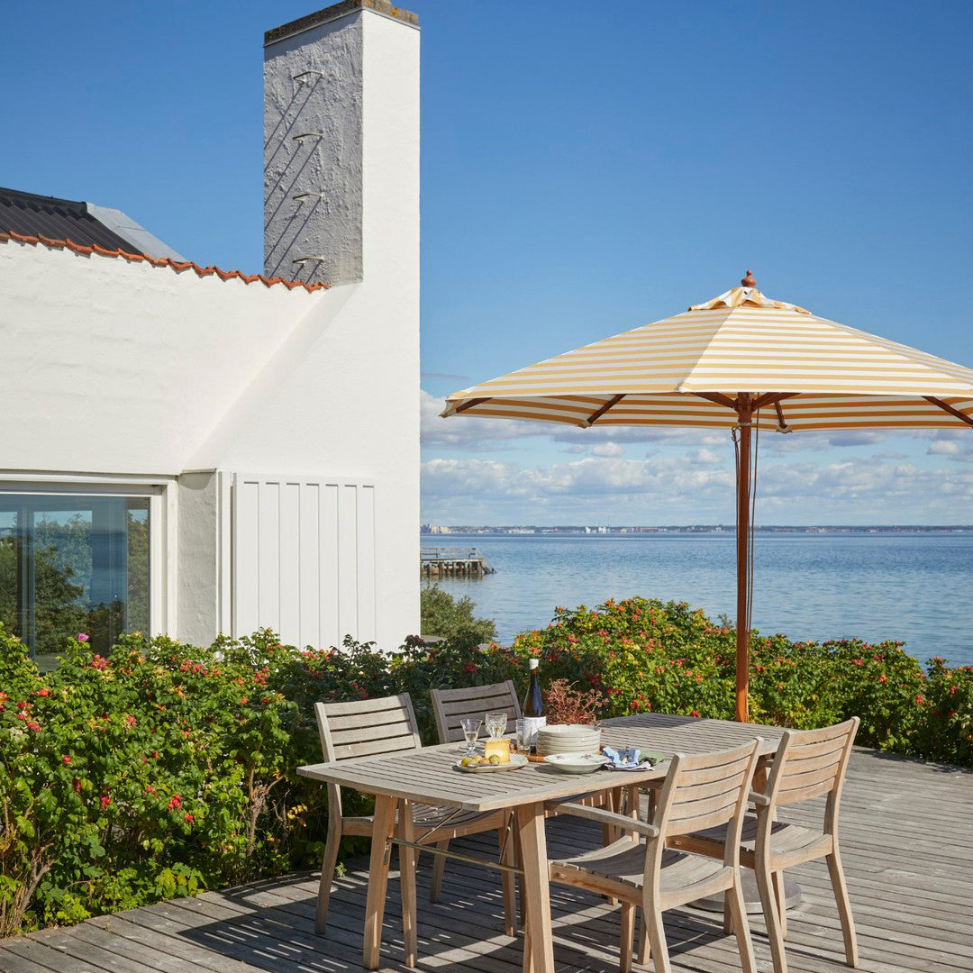 Outdoor dining set with natural wood tones, beige and white umbrella, vibrant green foliage, and blue water backdrop.