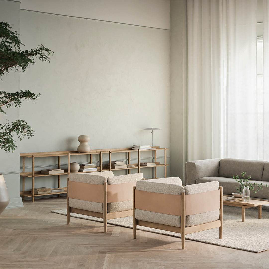 Pair of modern beige armchairs with light wood frames, set against a neutral backdrop and soft draped curtains.