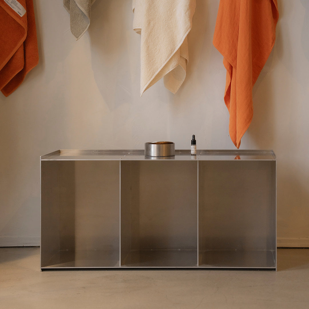 Sleek metallic cabinet with warm orange and neutral towels hanging above, highlighting modern minimalism and color contrast.