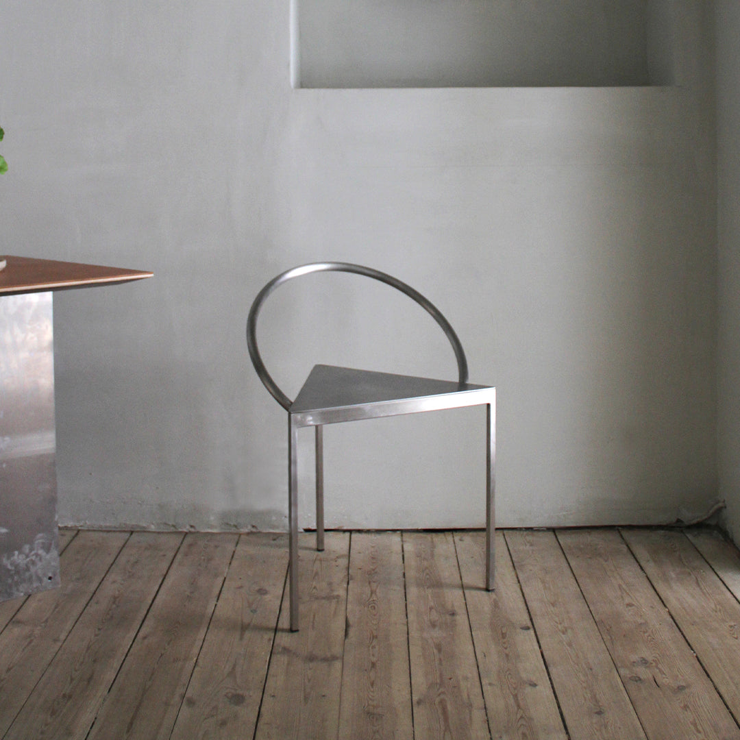 Minimalist metal chair with a sleek silver finish and unique triangular design against a soft gray background.