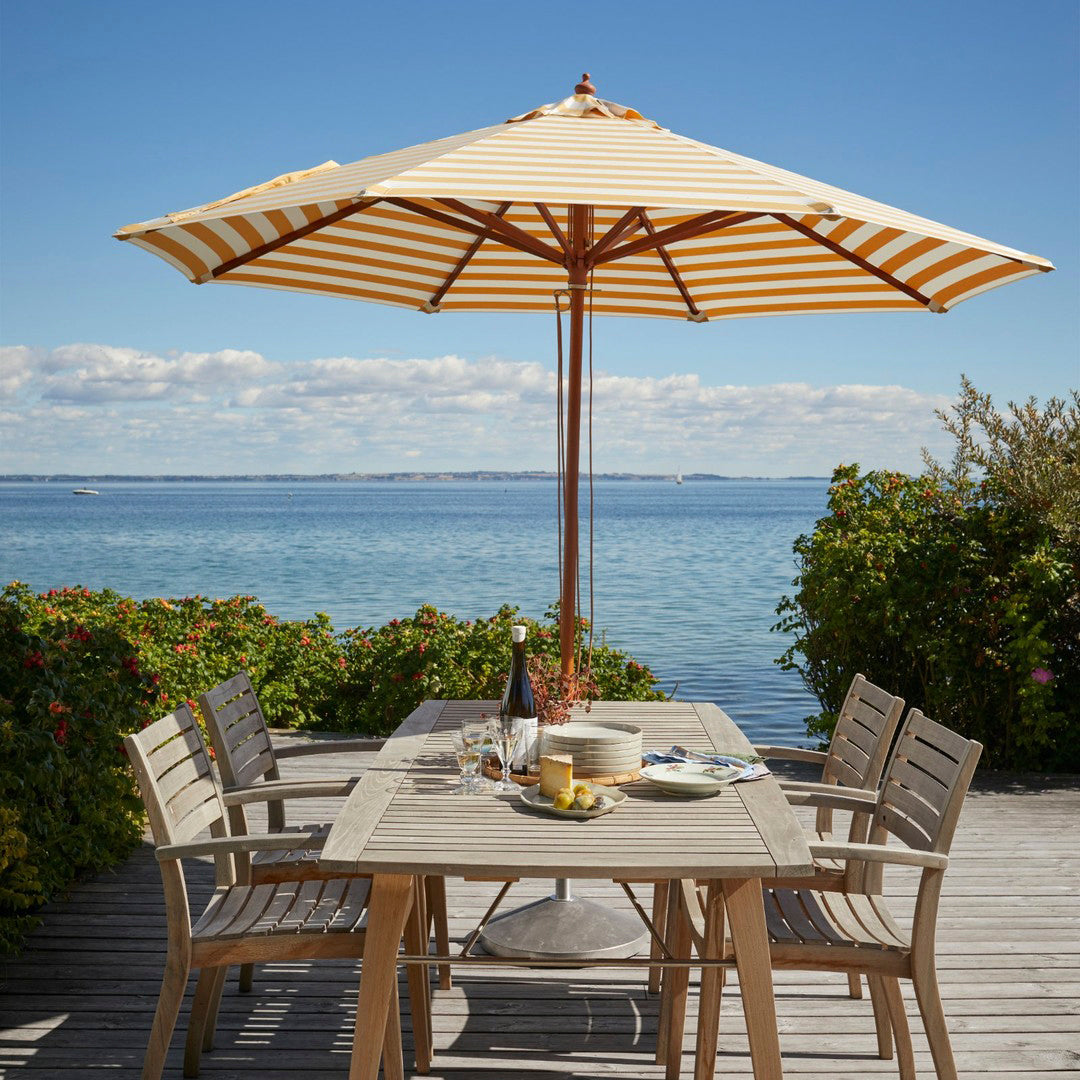 Yellow and white striped patio umbrella over a wooden table by the water, surrounded by greenery.