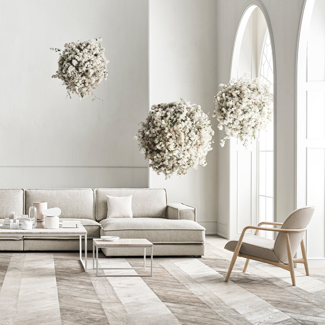 Light beige sofa in a minimalist room with white walls, patterned wooden floor, and hanging white floral arrangements.