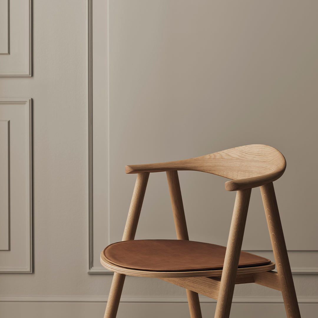 Natural wood chair with a light oak finish and a rich brown leather seat against a muted beige wall.