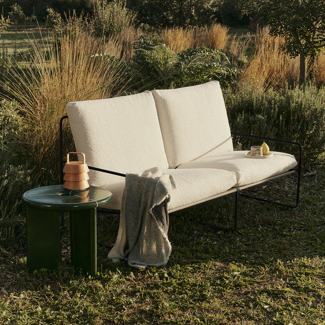 Cream outdoor sofa with sleek black frame, paired with a green side table, surrounded by lush greenery.