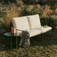 Cream outdoor sofa with sleek black frame, paired with a green side table, surrounded by lush greenery.