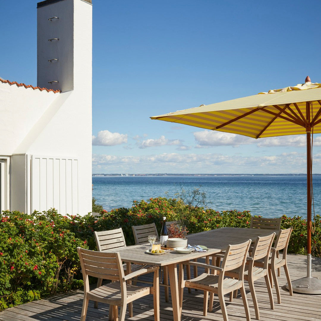"Bright yellow umbrella over wooden dining set, surrounded by lush greenery and a serene blue sea."