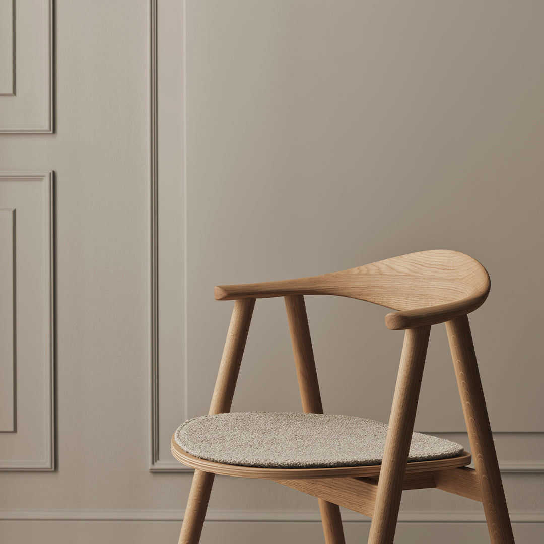 Light wood chair with a beige seat cushion, set against a neutral gray wall. Minimalist design with subtle textures.