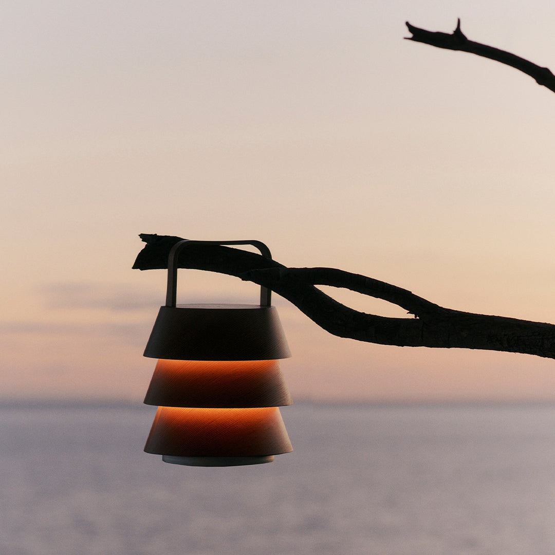 Stylish layered lamp in warm orange and sleek black, suspended from a branch against a serene sunset backdrop.