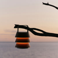Stylish layered lamp in warm orange and sleek black, suspended from a branch against a serene sunset backdrop.
