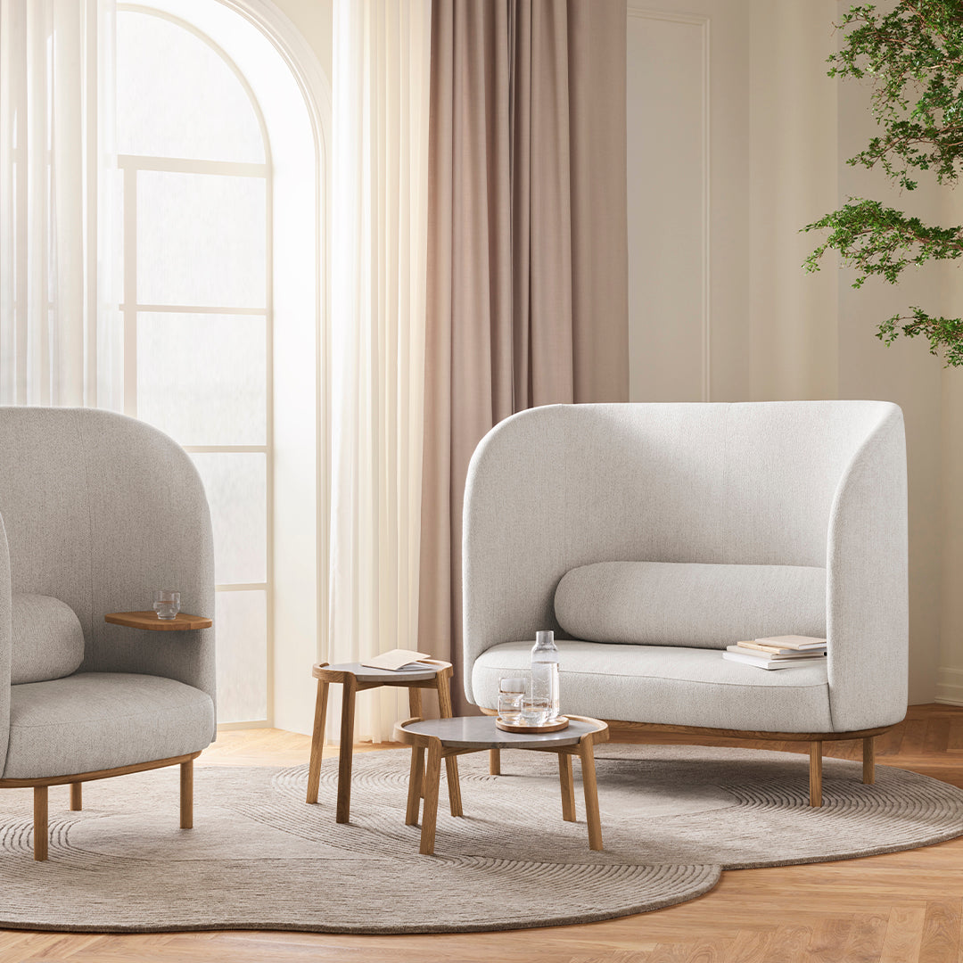 "Two soft gray upholstered chairs with rounded backs, paired with light wooden tables on a beige rug in a bright room."