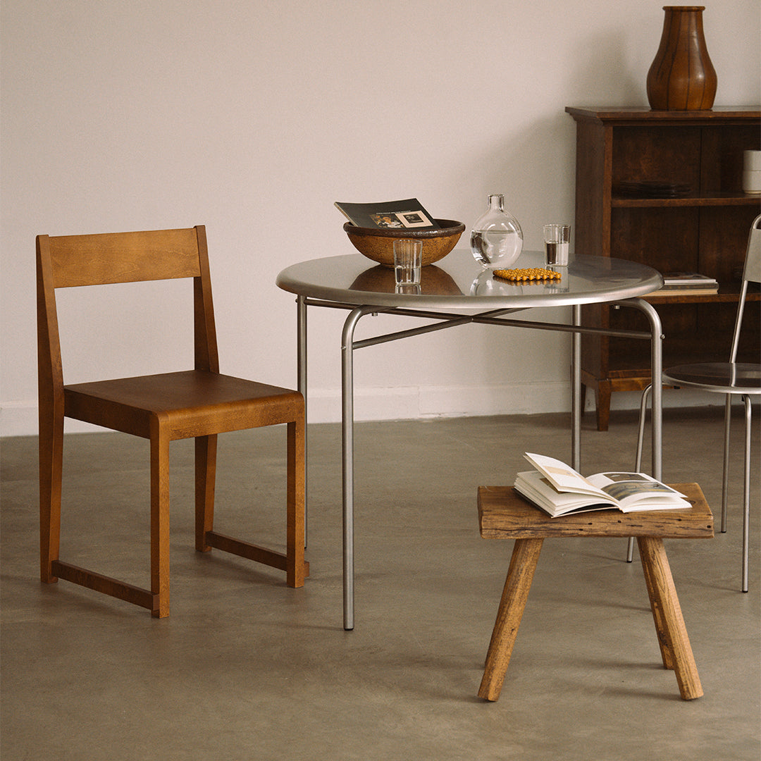 Modern wooden chair with a warm brown finish beside a round silver table; neutral backdrop enhances minimalist aesthetic.